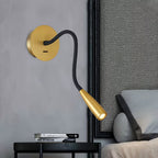 Gold Clérachic wall light with its black flexible arm bent downward over a bedside table and a grey bed pillow.