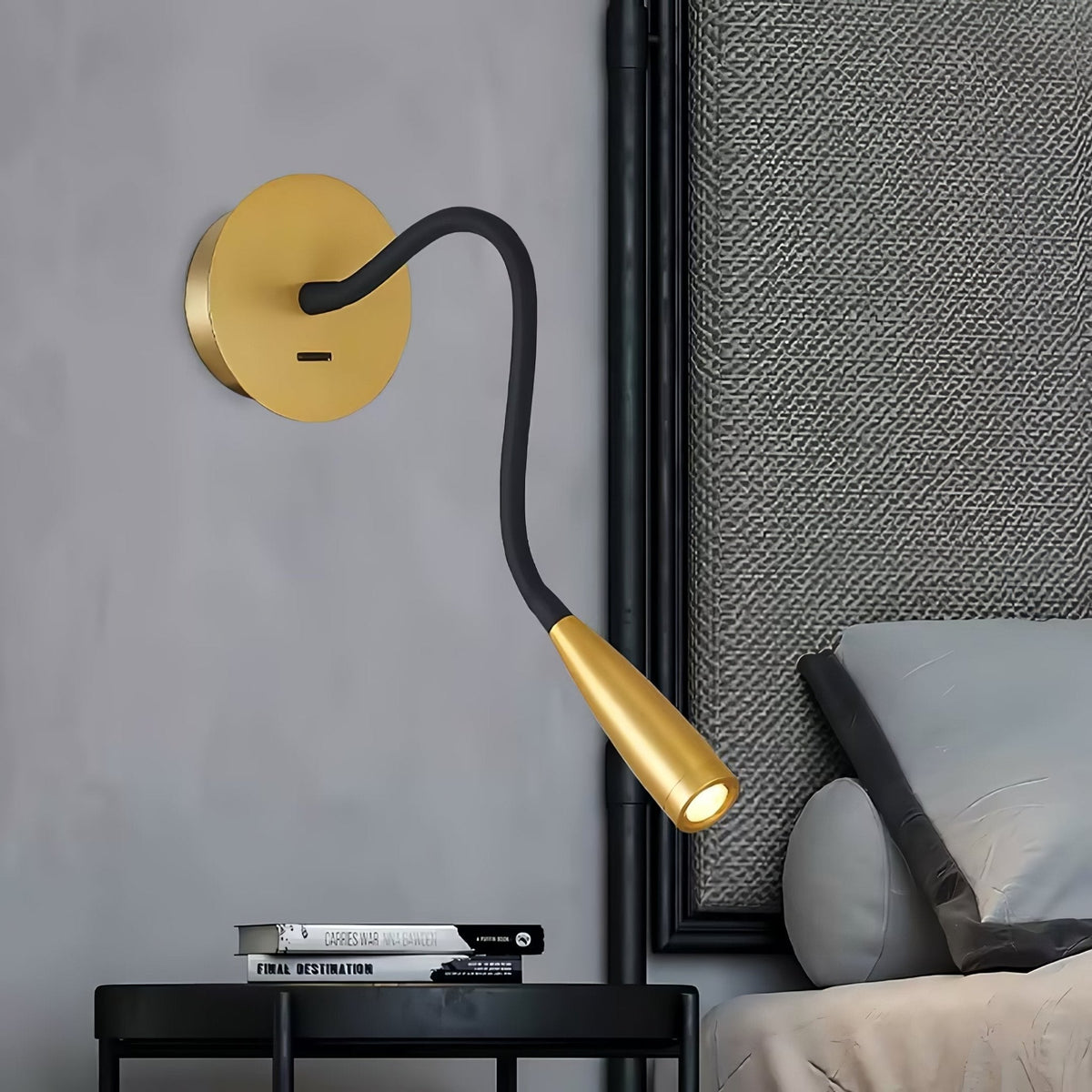 Gold Clérachic wall light with its black flexible arm bent downward over a bedside table and a grey bed pillow.