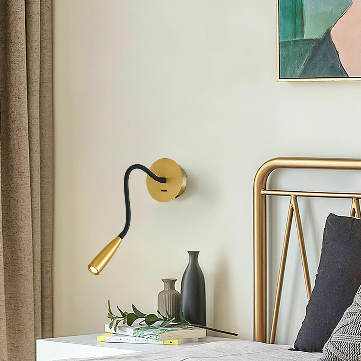 A gold Clérachic wall light with a curved black arm beside a bed with a brass headboard, over a nightstand with vases and books.