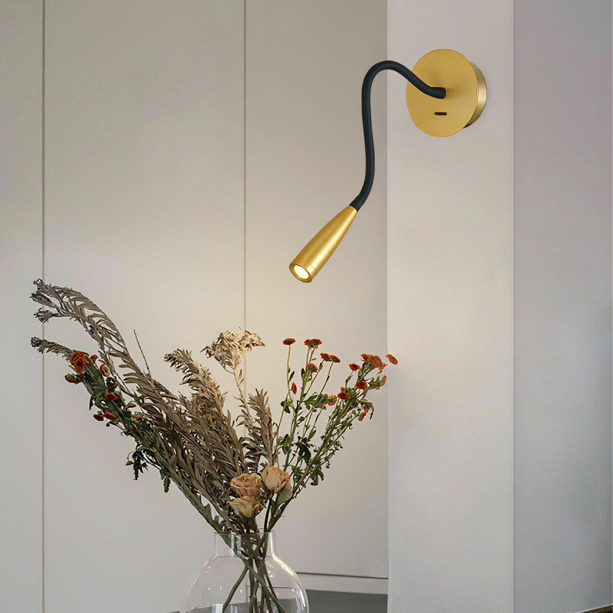 Clérachic wall light with a gold spot head bent downward, above a glass vase of dried grasses and red-orange flowers.
