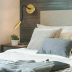 Gold Clérachic reading light on a dark textured wall above a bed with grey and cream cushions, its arm curved to point down.
