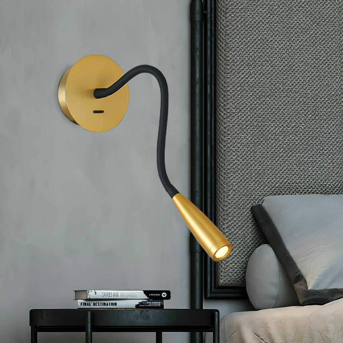 Gold Clérachic wall light with a black bendable arm and tapered gold spot head, set above a black side table and bed.