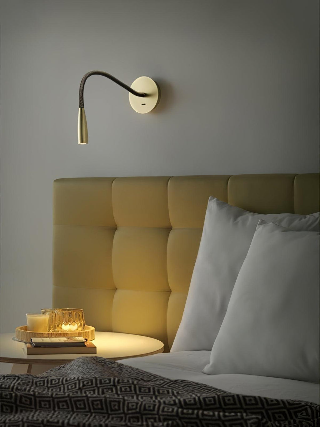 A gold Clérachic reading wall light above a tufted headboard, with a round tray of candles on a small bedside table below.
