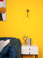 Small Clérachic wall light with a dark bronze finish on a bright yellow wall, above a white nightstand and a navy sofa.