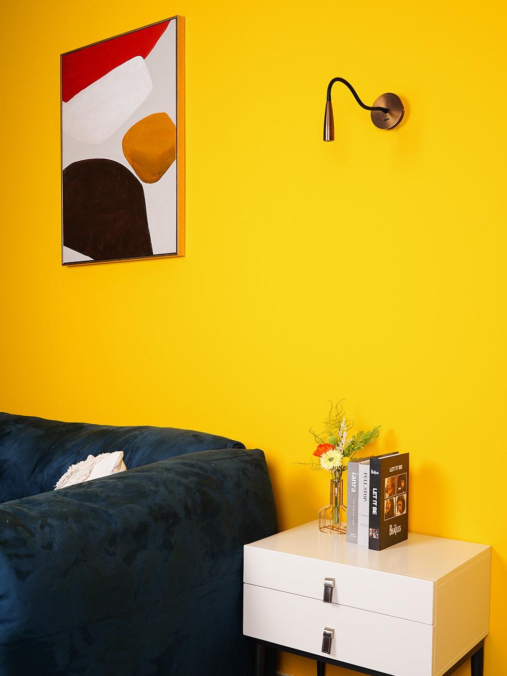Bronze-toned Clérachic wall light with its arm curved forward on a yellow wall, above a navy sofa and a white two-drawer nightstand.