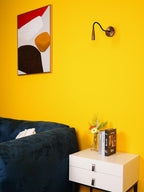 Bronze-toned Clérachic wall light with its arm curved forward on a yellow wall, above a navy sofa and a white two-drawer nightstand.
