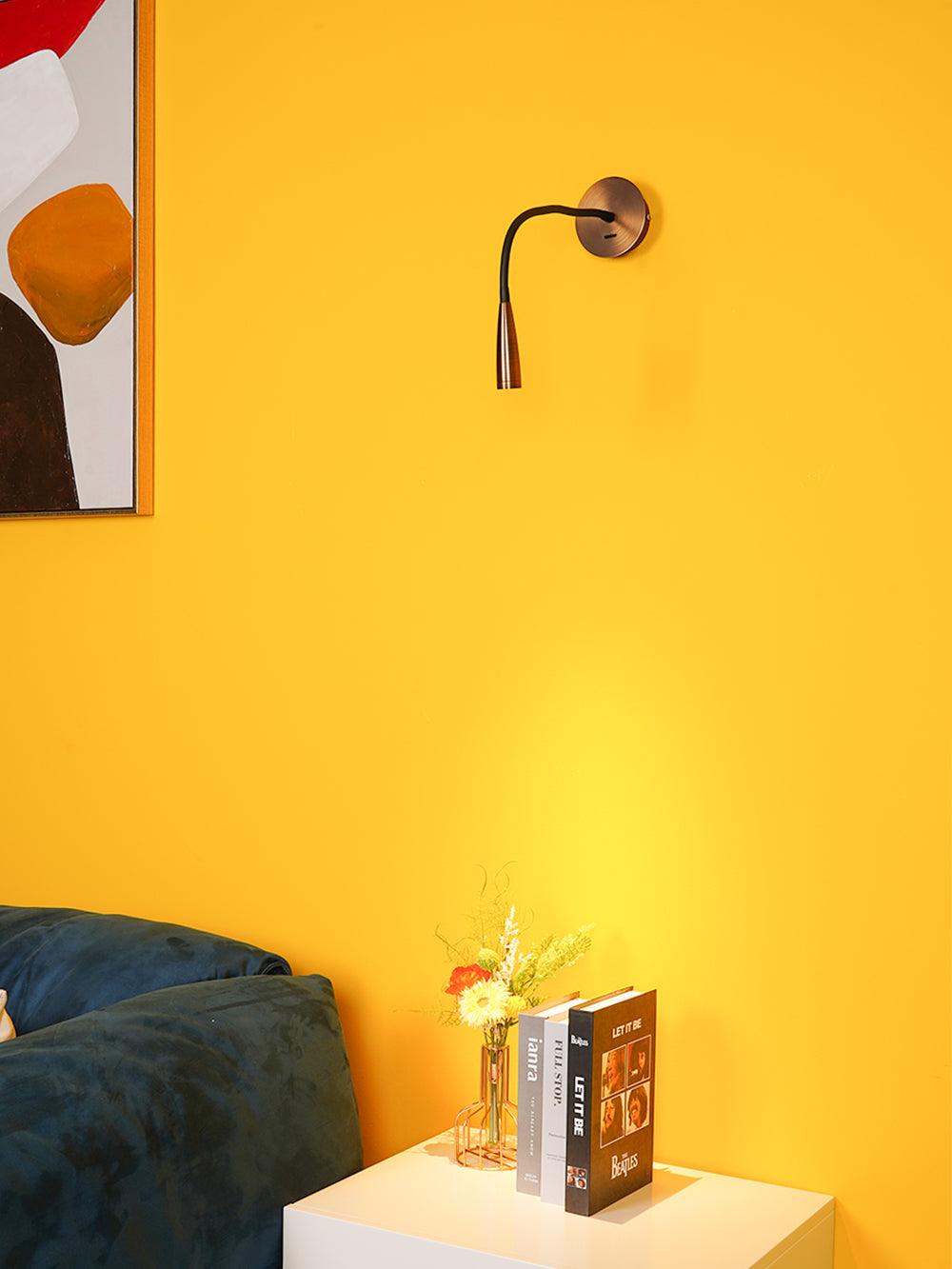 Dark bronze Clérachic wall light with a softly lit patch on the yellow wall below, above a white nightstand with flowers and books.