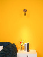 A distant view of this Clérachic wall light in bronze on a vivid yellow wall, with a warm light patch above a white nightstand.
