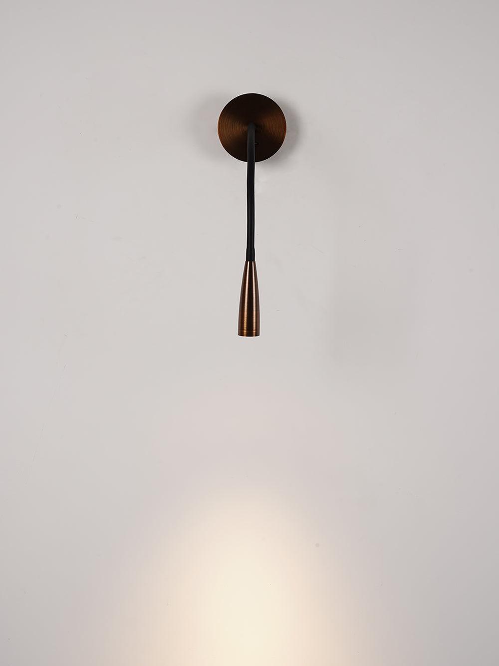 Lit Clérachic wall light in dark bronze with a straight, narrow black arm, throwing a soft pool of warm light down a pale wall.