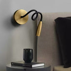 Gold Clérachic wall light with its black arm coiled in a loop, beside a bed with a grey headboard, above a round table with a book and cup.