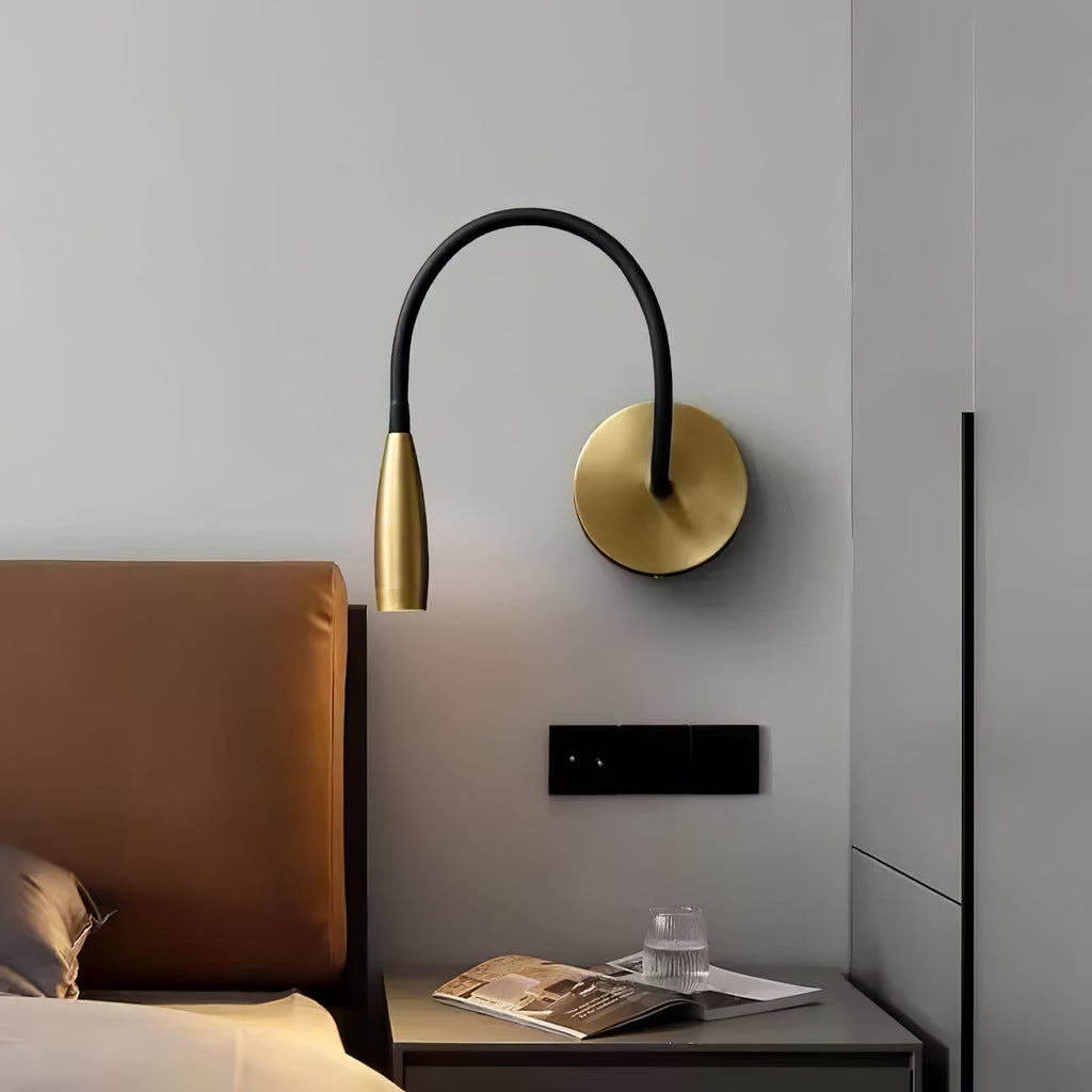 Gold Clérachic wall light with a high black arched arm, above a bedside table with a magazine, beside a tan leather headboard.