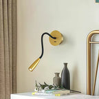 Gold Clérachic wall light on a cream wall beside a brass bed frame, above a white nightstand with two vases, a leafy stem and books.
