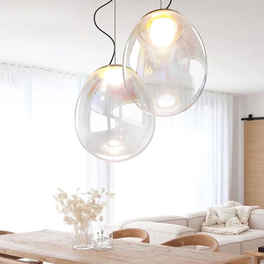 Two clear glass teardrop pendants from Clérachic hang at different heights over a wooden dining table, with a sofa and sheer curtains behind.