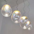 Seen from below on a grey wall, four lit Clérachic glass pendants step down on wavy dark cords, each with a warm glowing bulb.