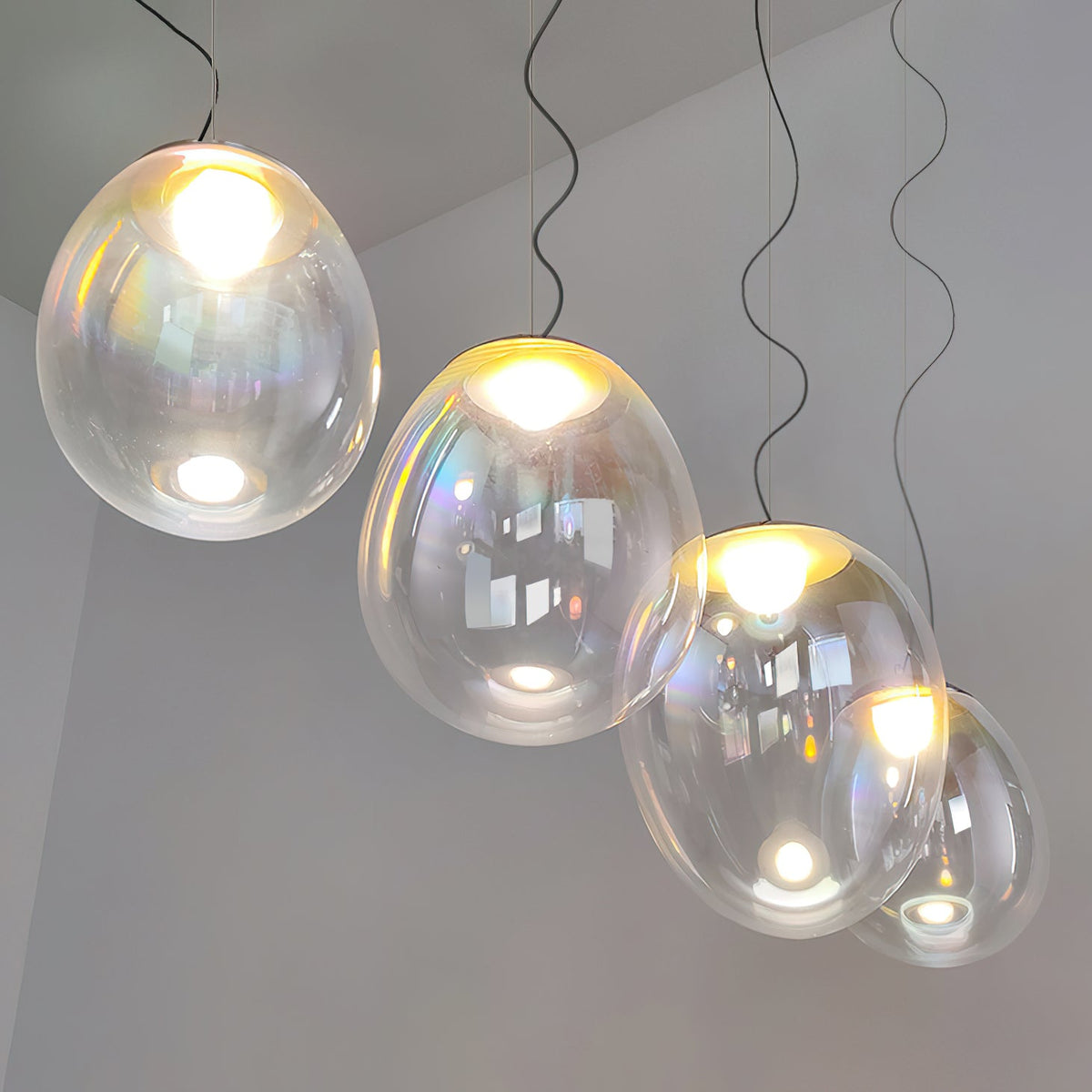 Seen from below on a grey wall, four lit Clérachic glass pendants step down on wavy dark cords, each with a warm glowing bulb.