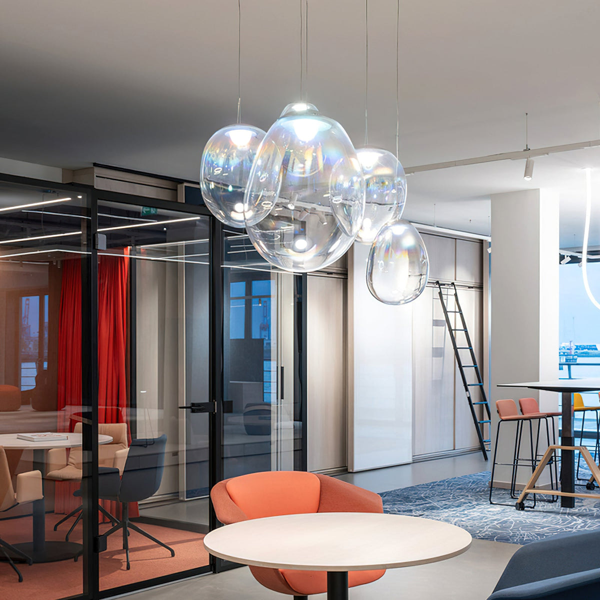 A tight group of Clérachic clear glass pendants hangs in a bright open office with glass partitions, a red curtain and a round white table.