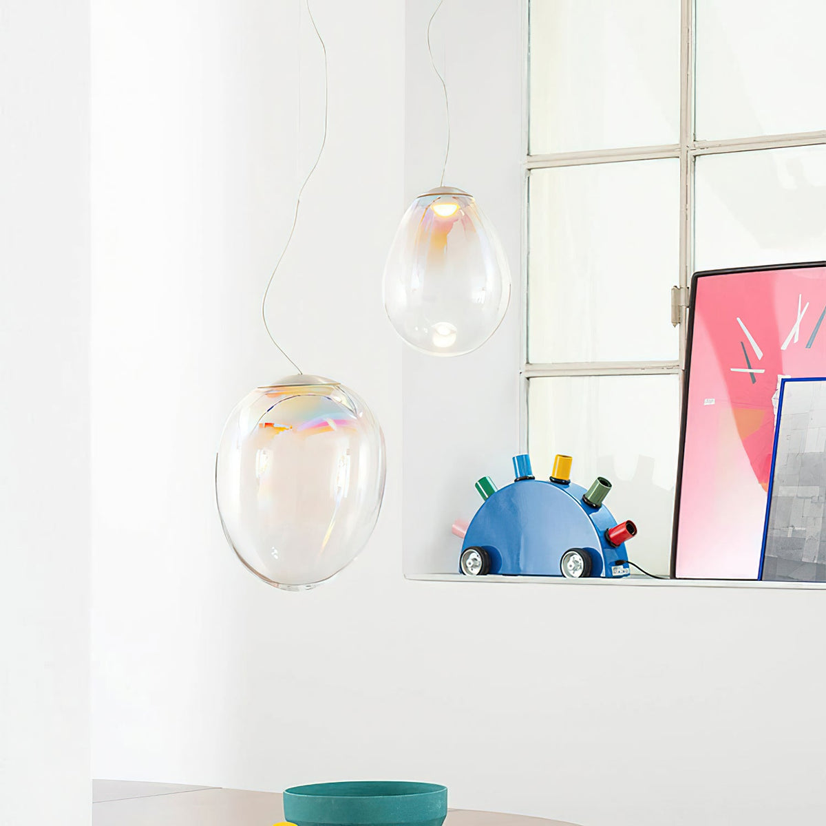 Two Clérachic glass teardrop pendants hang beside a grid window, above a table with a teal bowl and a blue shelf ornament.