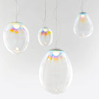 Four Clérachic glass teardrop pendants of varied size glow against a light backdrop, each with a rainbow sheen around its top cap.