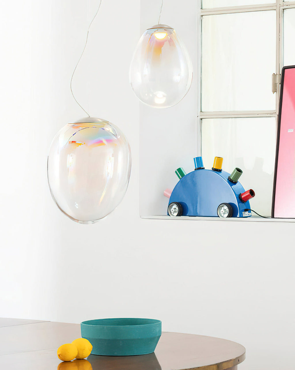 Lower view of this Clérachic glass pendant pair in a white room, with a blue ornament on a shelf and a pink poster beside a window.