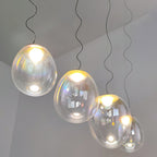 Four lit Clérachic glass pendants step down on dark cords in a pale room, each shade clear with a faint rainbow tint and a bright bulb.