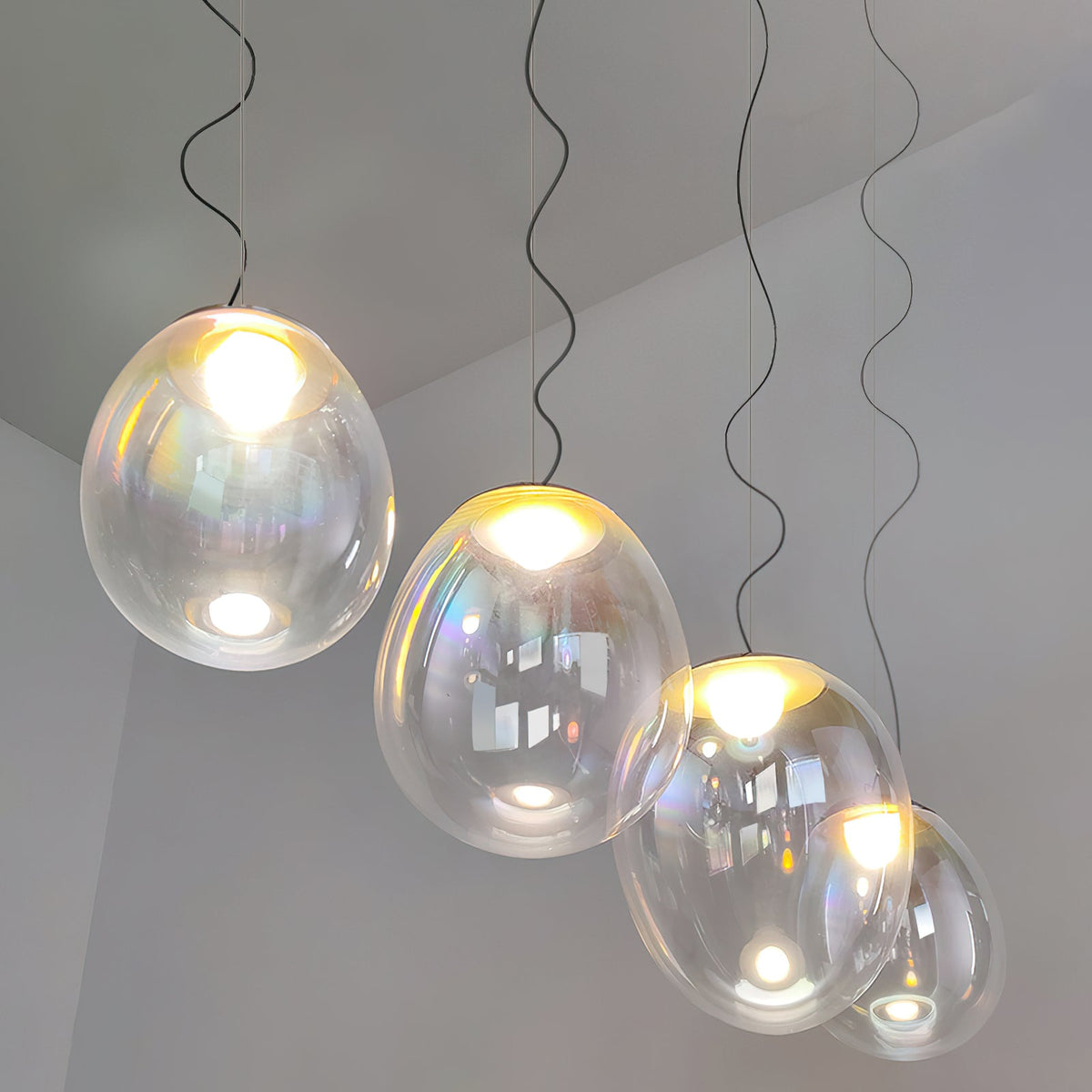 Four lit Clérachic glass pendants step down on dark cords in a pale room, each shade clear with a faint rainbow tint and a bright bulb.