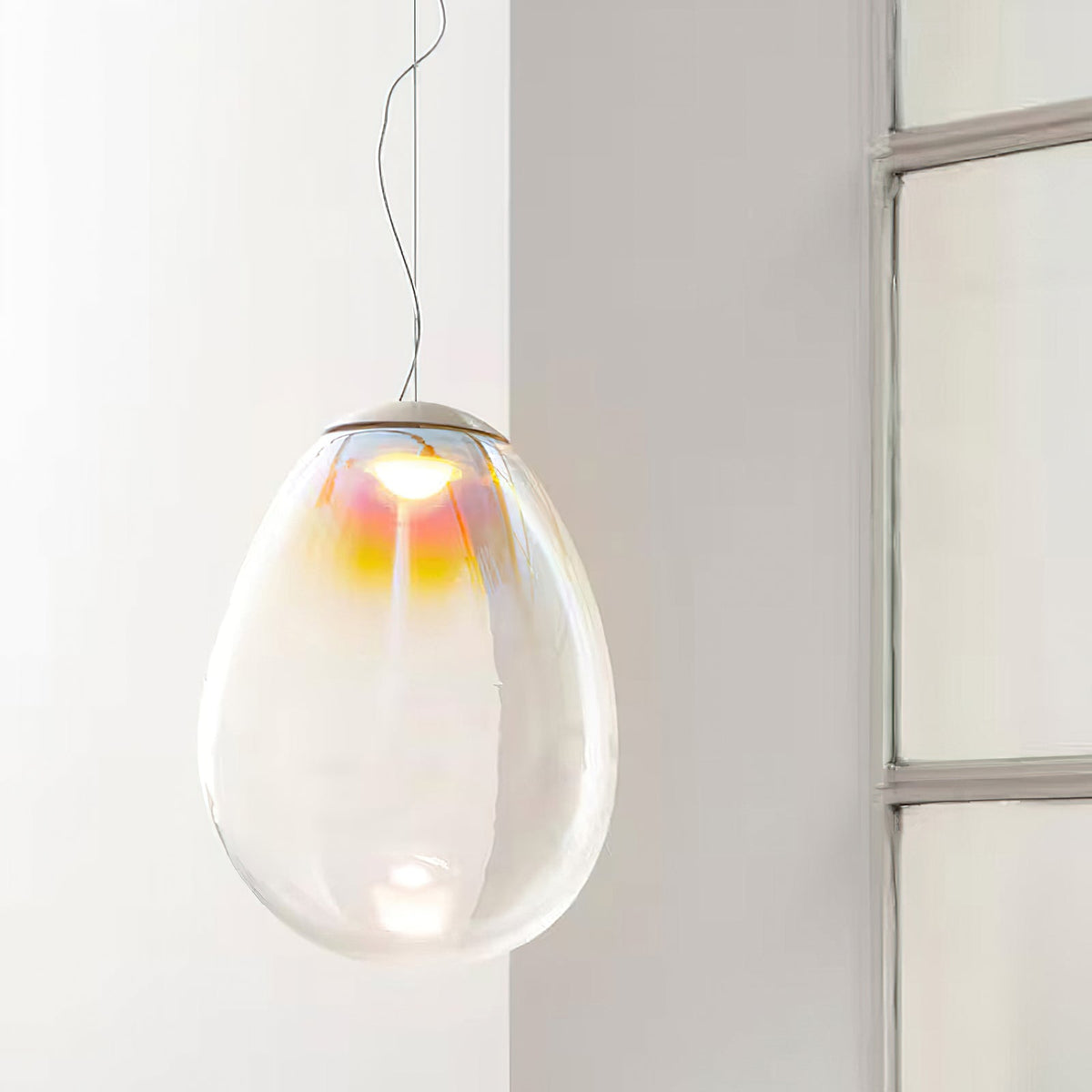 This Clérachic glass pendant hangs on a thin cord near a window, its clear egg-shaped shade tinted gold and pink at the top.