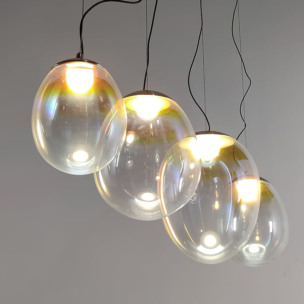 Four Clérachic glass pendants hang lit at varied heights on dark cords against a grey wall, their shades showing soft rainbow reflections.