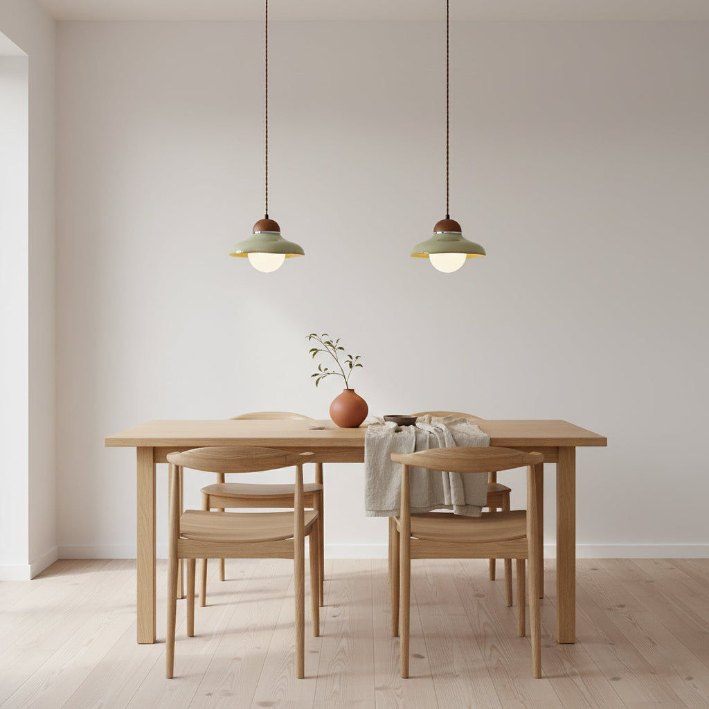 Two small Clérachic green pendants hang over a pale wooden dining table with four chairs, against a plain wall.