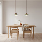 Two small Clérachic green pendants hang over a pale wooden dining table with four chairs, against a plain wall.