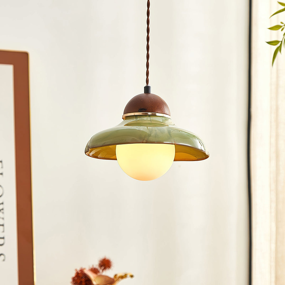 Clérachic green ceramic pendant with a lit globe, next to a framed print and a window with a cream curtain.