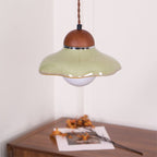 Wavy-edged green ceramic Clérachic pendant with a walnut cap, hanging in a corner above a wooden sideboard.