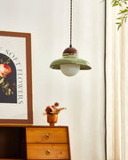 A small Clérachic green pendant hangs above a wooden chest of drawers, beside a framed floral print and a curtain.