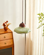 Clérachic green ceramic pendant seen from beside a wooden drawer unit, with a cream curtain and a leafy stem behind.