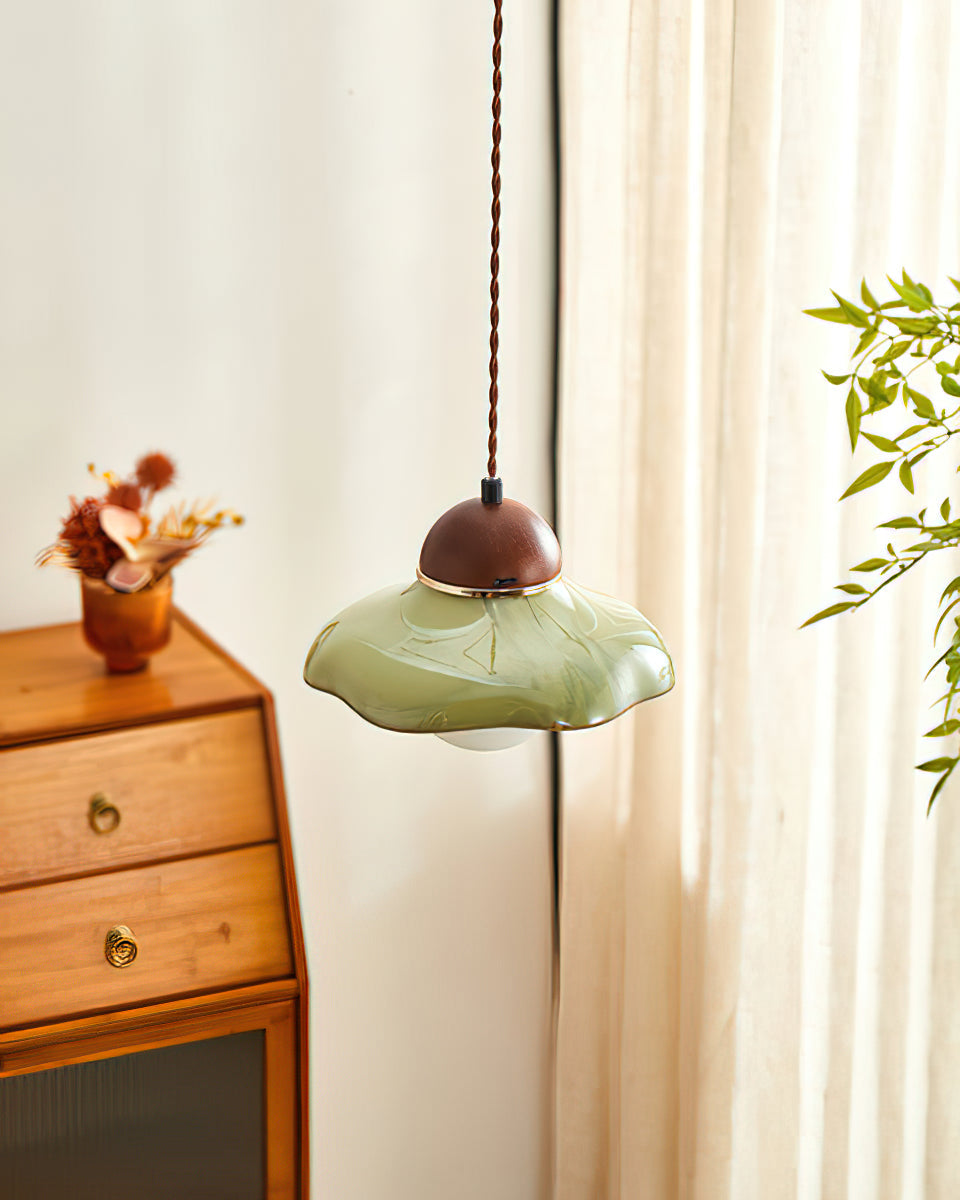 Clérachic green ceramic pendant seen from beside a wooden drawer unit, with a cream curtain and a leafy stem behind.