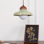 Clérachic green pendant with a lit white globe, hanging above a framed gold floral print on a dark wooden surface.