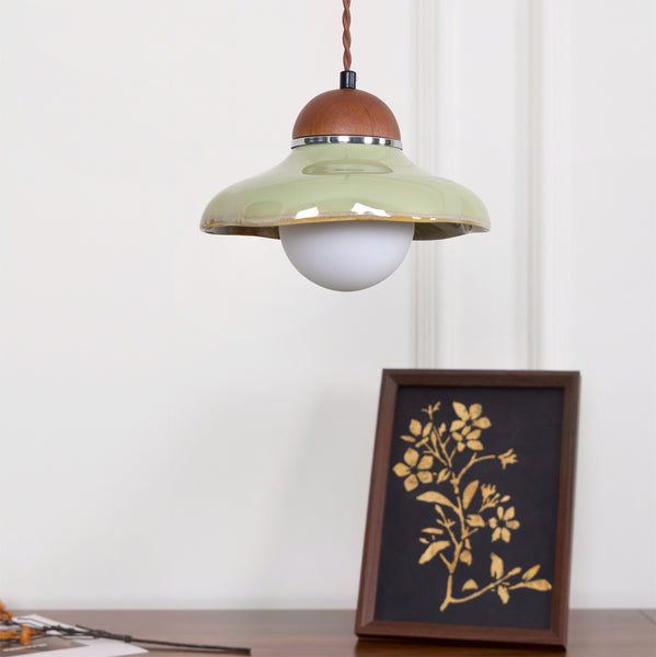 Clérachic green pendant with a lit white globe, hanging above a framed gold floral print on a dark wooden surface.