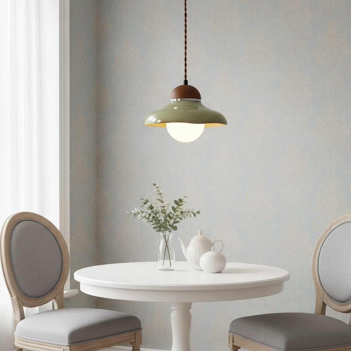A Clérachic green ceramic pendant above a round white table with a vase and teapots, between two grey cushioned chairs.