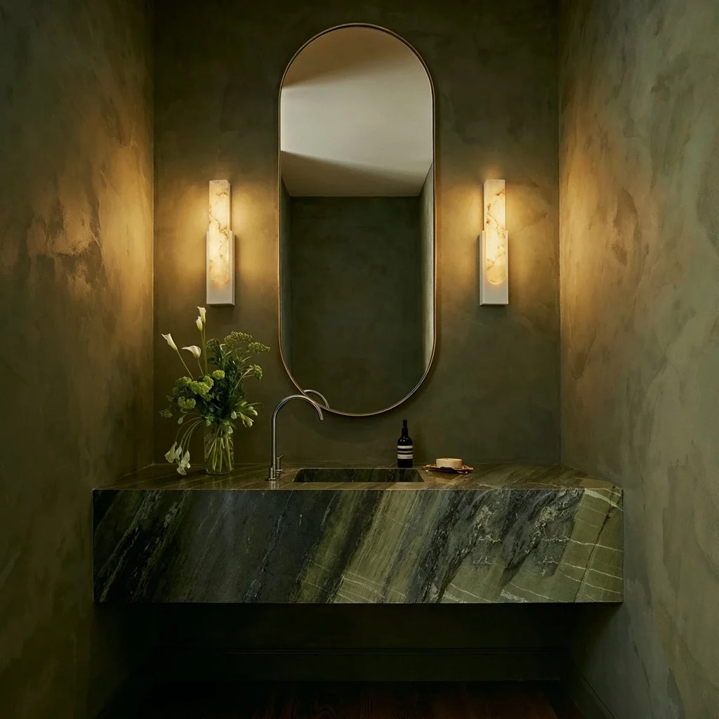 Pair of Clérachic white-framed alabaster sconces flanking an oval mirror above a green marble bathroom vanity.