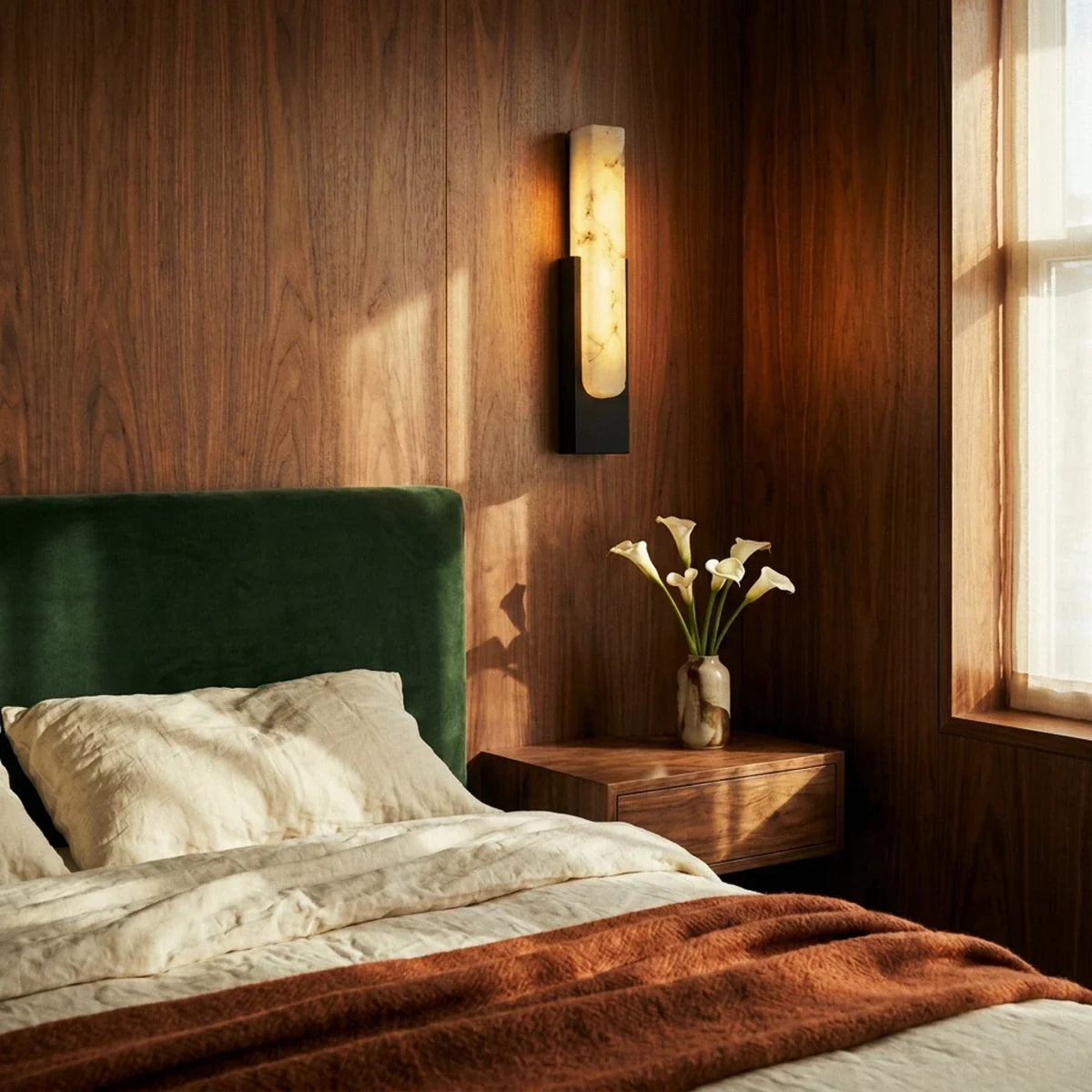 Clérachic black-framed alabaster sconce glowing beside a bed with a green velvet headboard in a walnut bedroom.