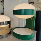 Close, low view of a Clérachic table lamp with a glowing dome and a green metal body, a chrome lamp visible behind it.