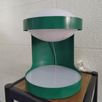 Close, straight-on view of this Clérachic table lamp's green metal body, its pale dome and a thin white cord, on a small wood-top stand.