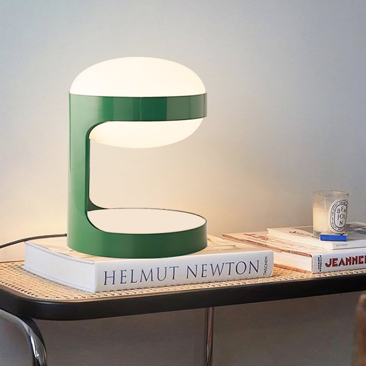 Lit Clérachic table lamp on a stack of large books beside a candle, on a cane-top black side table, warm light on the wall.