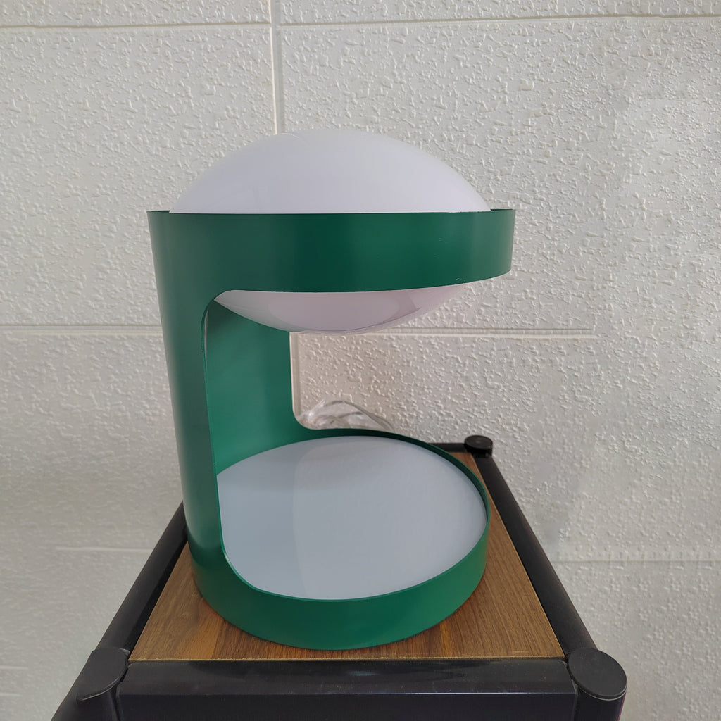 Side view of this Clérachic table lamp on a wood-top stand against a tiled wall, the opening of its green body facing left.