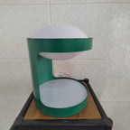 Side view of this Clérachic table lamp on a wood-top stand against a tiled wall, the opening of its green body facing left.