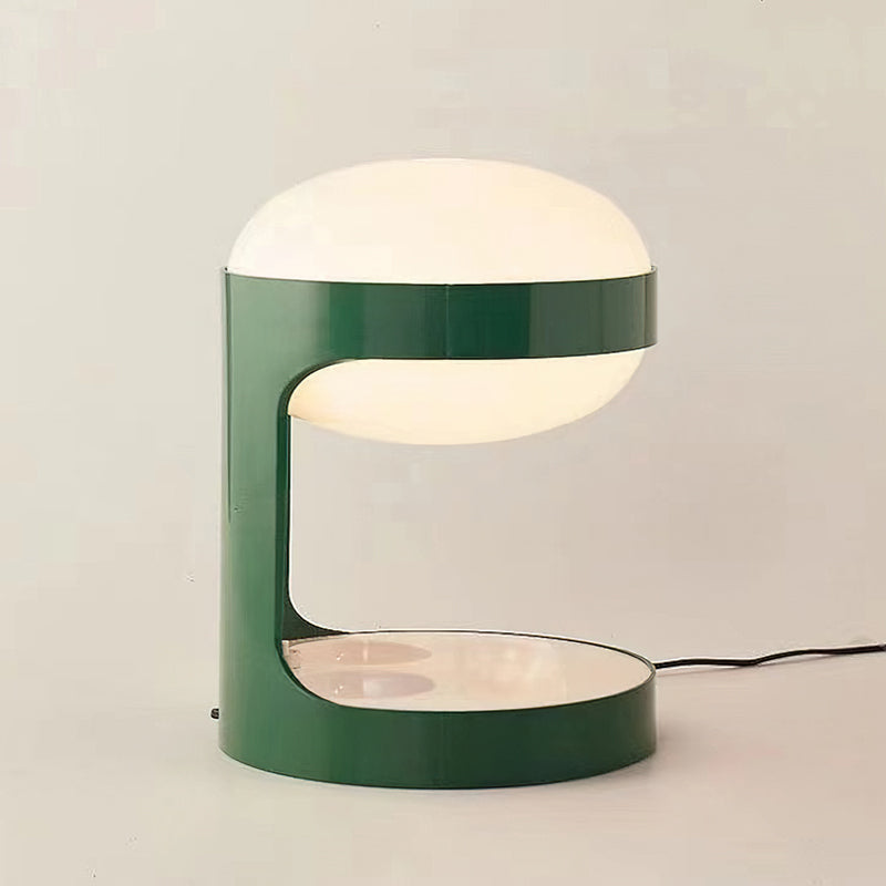 Clérachic table lamp lit on a soft beige backdrop, its C-shaped green body holding the glowing dome, with a black cord to the right.
