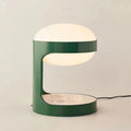 Clérachic table lamp lit on a soft beige backdrop, its C-shaped green body holding the glowing dome, with a black cord to the right.