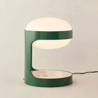 Clérachic table lamp lit on a soft beige backdrop, its C-shaped green body holding the glowing dome, with a black cord to the right.