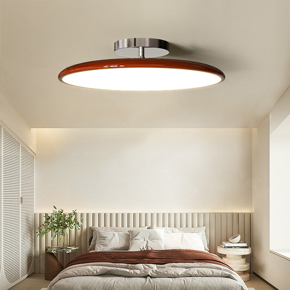 Clérachic deep red LED disc ceiling light lit above a bed with warm rust-toned bedding and slatted headboard.