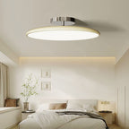 Clérachic cream LED disc ceiling light casting a warm glow in a bedroom with a lit nightstand lamp nearby.