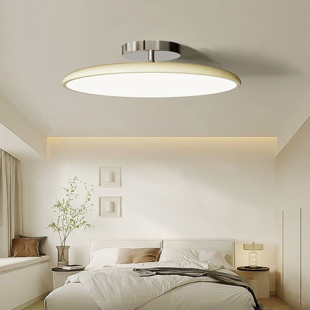 Clérachic cream LED disc ceiling light casting a warm glow in a bedroom with a lit nightstand lamp nearby.